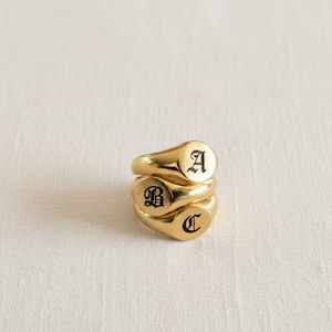 May include: Three gold-colored signet rings stacked on top of each other. Each ring has a rounded face with a black letter: A, B, and C. The rings are on a white, textured surface.