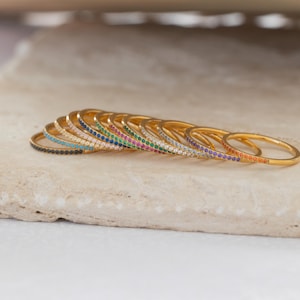 May include: A collection of gold-colored rings, each adorned with a row of colorful gemstones. The gemstones include black, turquoise, white, blue, green, pink, purple, and orange. The rings are displayed on a light-colored, textured surface.