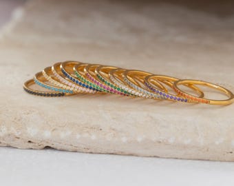 Pode incluir: Uma coleção de anéis dourados, cada um adornado com uma fileira de pedras preciosas coloridas. As pedras preciosas incluem preto, turquesa, branco, azul, verde, rosa, roxo e laranja. Os anéis são exibidos em uma superfície clara e texturizada.