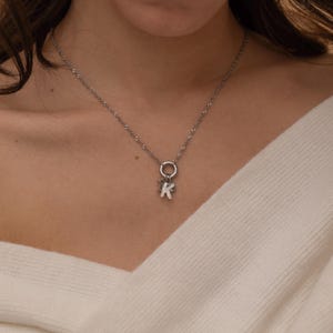 Könnte beinhalten: Eine silberfarbene Halskette mit einer feinen Kette und einem runden Anhängerring. An dem Ring befindet sich ein kleiner, strukturierter Anhänger in Form des Großbuchstabens K. Die Kette liegt auf einem weißen Kleidungsstück mit V-Ausschnitt.