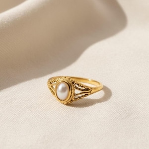 May include: A gold-colored ring with an oval, white pearl center stone. The ring features an ornate, filigree design around the pearl and along the band. The ring is set against a neutral, off-white background.