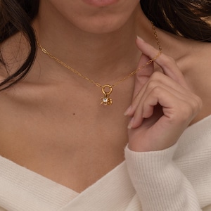 May include: A delicate gold necklace with a small pendant featuring a clear gemstone and a cluster of smaller gold elements. The necklace is worn on a person, and the chain has a paperclip style.