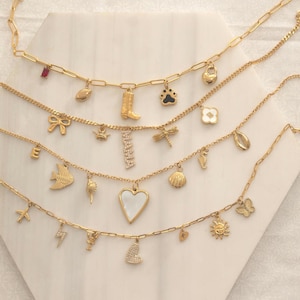 Puede incluir: Collares dorados con colgantes variados, incluyendo un corazón, una mariposa y un rayo. Los colgantes cuelgan de cadenas delicadas, creando un efecto de capas. Los collares se exhiben sobre una superficie blanca.