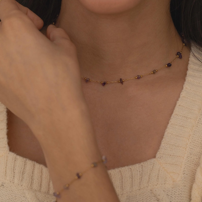 May include: Delicate gold necklace and bracelet set. The necklace features small, purple gemstones spaced along a fine gold chain. The bracelet mirrors the necklace design, creating a coordinated jewelry set. The jewelry is worn on a person.