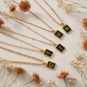 Op de afbeelding: Goudkleurige kettingen met zwarte rechthoekige hangers, elk met een andere letter in witte letters. De kettingen zijn gerangschikt op een wit oppervlak met gedroogde bloemen, wat een gepersonaliseerde sieradencollectie laat zien.