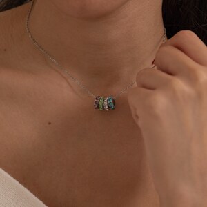 May include: A silver necklace featuring a series of colorful, gem-studded rings. The rings are arranged in a cluster, showcasing a spectrum of colors including green, purple, blue, and pink. The necklace is delicate and appears to be worn close to the neck.