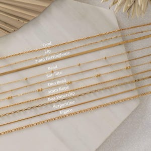 May include: A collection of gold-colored necklaces displayed on a white surface. The necklaces include various styles such as oval, lip, herringbone, bead satellite, duo bead, 3mm rope, box paperclip, serpentine, and figaro. The names of the necklace styles are printed on the surface.