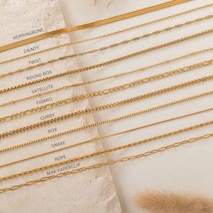 May include: A collection of gold-colored necklaces in various styles, including herringbone, dainty, twist, round box, satellite, figaro, curby, box, snake, rope, and mini paperclip. The necklaces are displayed on a light-colored surface.