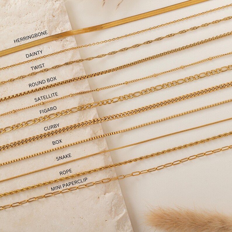 May include: A collection of gold-colored necklaces in various styles, including herringbone, dainty, twist, round box, satellite, figaro, curby, box, snake, rope, and mini paperclip. The necklaces are displayed on a light-colored surface.