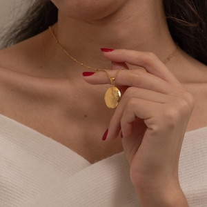 May include: A gold-colored locket necklace with a delicate chain. The oval locket features an embossed floral design. The necklace is being held by a person with red nail polish, and is set against a white sweater.