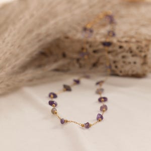 May include: A delicate gold necklace featuring small, irregularly shaped amethyst stones. The necklace is strung on a fine gold chain with small gold beads interspersed between the amethyst stones. The necklace is displayed on a white surface.