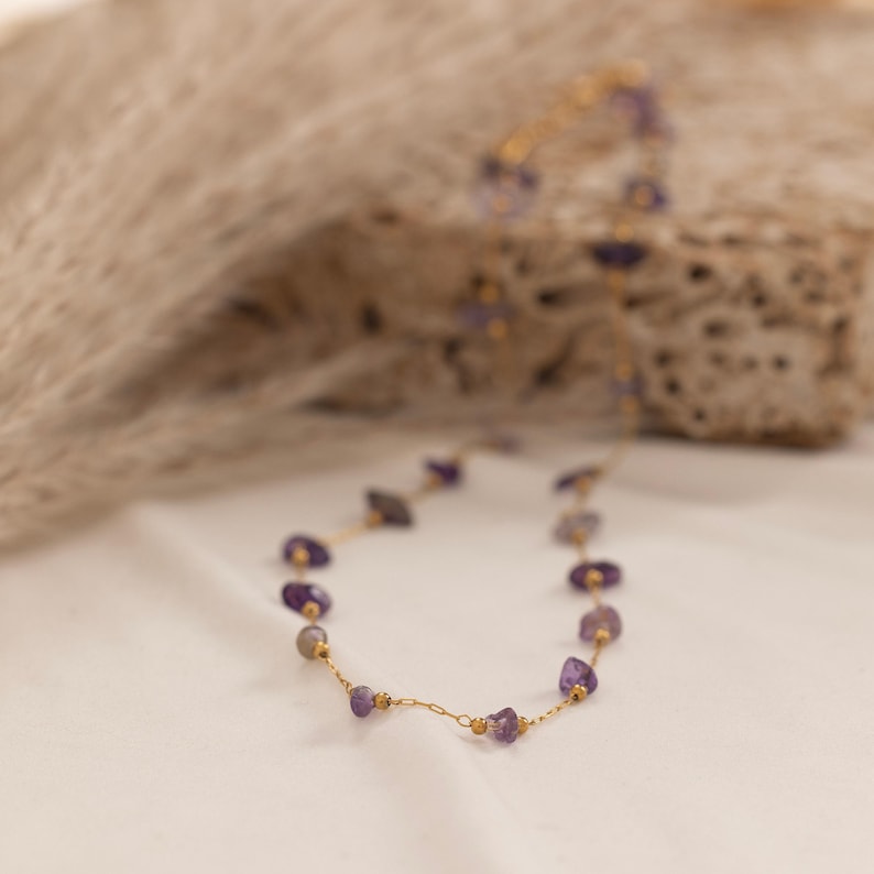 May include: A delicate gold necklace featuring small, irregularly shaped amethyst stones. The necklace is strung on a fine gold chain with small gold beads interspersed between the amethyst stones. The necklace is displayed on a white surface.