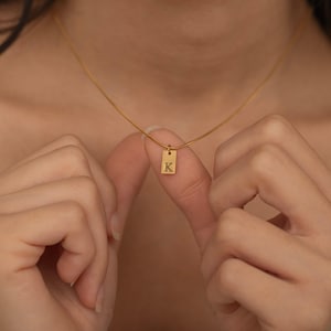 May include: A delicate gold-colored necklace featuring a rectangular pendant with the letter "K" engraved on it. The pendant hangs from a thin chain. The necklace is being held up by a person's fingers.