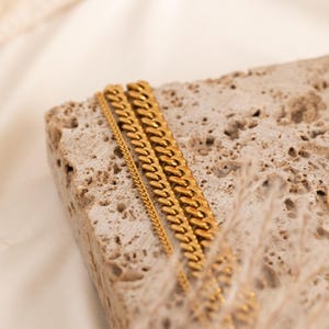 May include: Three gold-colored chain necklaces of varying widths are displayed on a textured, light-brown stone. The chains have a curb link design, with the links lying flat. The background is a soft, off-white color.