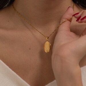 May include: A gold-colored oval locket pendant necklace. The pendant hangs from a delicate chain. The locket is being held by a person with red nail polish. The necklace is a piece of jewelry.