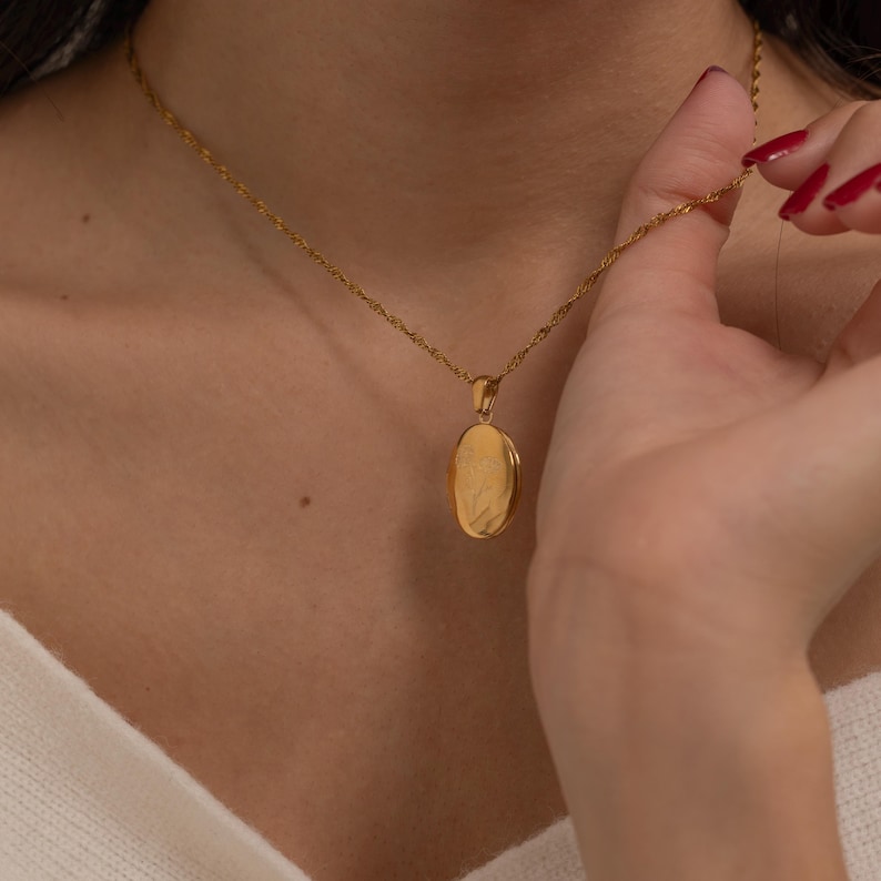 May include: A gold-colored oval locket pendant necklace. The pendant hangs from a delicate chain. The locket is being held by a person with red nail polish. The necklace is a piece of jewelry.