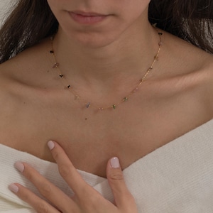 May include: Delicate gold necklace featuring small, colorful gemstones. The necklace is strung with a fine gold chain and showcases a variety of natural stone colors, including green, purple, and black. The necklace is a simple yet elegant piece of jewelry.