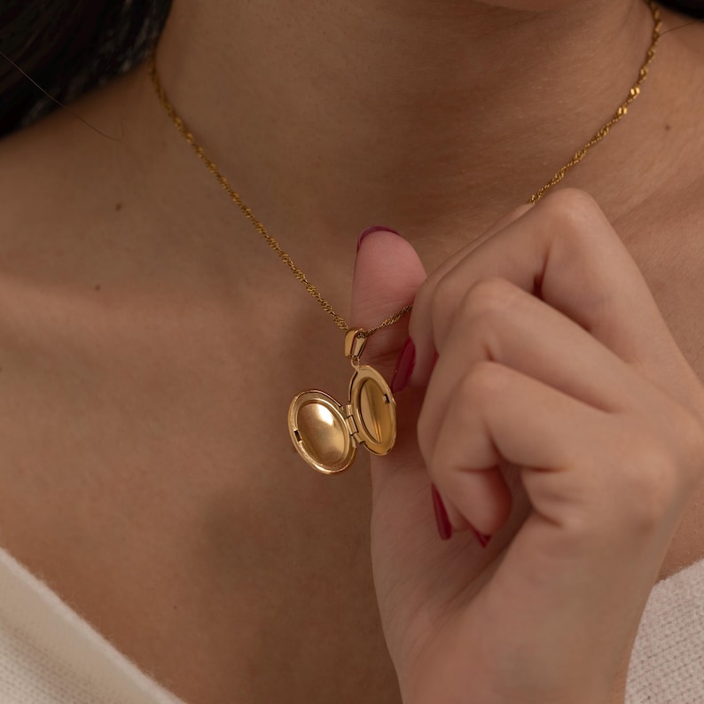 May include: A gold-colored oval locket necklace with an open pendant, revealing a mirrored interior. The necklace is on a delicate chain. The locket is being held open by a hand with red nail polish.