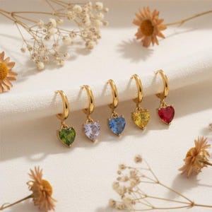 May include: Gold hoop earrings with heart-shaped gemstones in green, purple, blue, yellow, and red. The earrings are displayed on a white cloth with dried flowers in the background. The gemstones are approximately 0.5 inches in size.