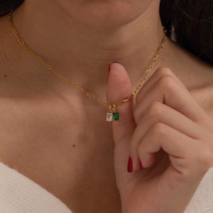 Könnte beinhalten: Goldfarbene Halskette mit feiner Kette und zwei rechteckigen Anhängern. Ein Anhänger hat einen klaren Stein, der andere einen grünen Stein. Die Halskette wird um den Hals getragen.