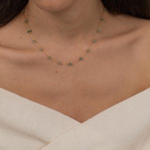 May include: A delicate gold necklace featuring small, irregularly shaped green gemstones. The necklace is worn around the neck, resting on a white top. The gemstones are evenly spaced along the gold chain.