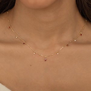 May include: A gold chain necklace featuring a series of small, dangling charms. The charms alternate between tiny, clear, round stones and small, deep red, teardrop-shaped stones, all evenly spaced along the delicate, thin gold metal chain.