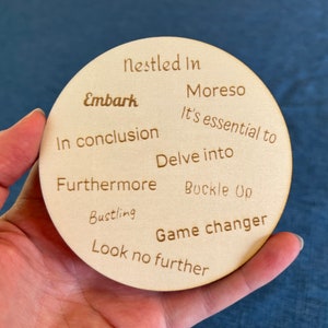 May include: A round, light-colored wooden coaster with various phrases etched into its surface, including "Embark," "In conclusion," and "Game changer." The coaster is held in a hand against a blue background.