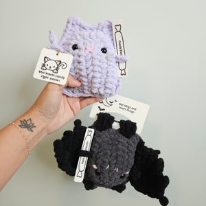 May include: Handmade crochet cat and bat plush toys. The lavender cat has a pink nose and the text "Who wants candy right meow?". The dark gray bat has black wings and the text "Bat wings and sweet things". Both have candy-shaped tags.