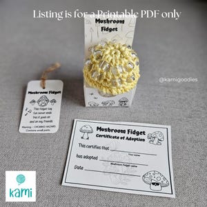 May include: A yellow crocheted mushroom fidget toy with silver accents, displayed in a small box. The image includes a tag and a certificate of adoption, both with mushroom graphics and text. The text on the tag reads "The fidget toy fun never ends".
