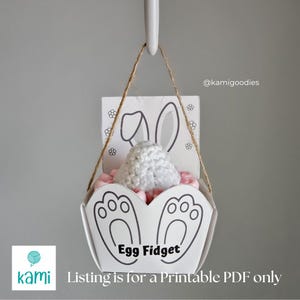 Könnte beinhalten: Ein weißer Osterkorb aus Papier mit Hasenmuster, der ein weißes gehäkeltes Ei und rosa Füllung enthält. Der Korb hat einen Griff und den Text "Egg Fidget". Der Text "Listing is for a Printable PDF only" ist ebenfalls sichtbar.