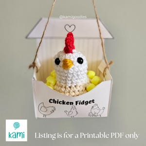 May include: A white crocheted chicken with a red comb and black eyes sits in a white box with the text "Chicken Fidget" and a drawing of three chickens. The box is hanging from a string.