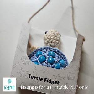 Könnte beinhalten: Ein gehäkeltes Schildkröten-Fidget-Spielzeug mit blauen Perlen. Die Schildkröte befindet sich in einer weißen Schachtel mit einem Meeres-Muster und dem Text "Turtle Fidget" und "Listing is for a Printable PDF only".