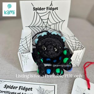 May include: A black crocheted spider fidget toy with blue and green accents, presented in a white box with a spiderweb design. The box reads "Spider Fidget". The listing is for a printable PDF.