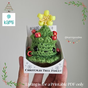 May include: A green crocheted Christmas tree fidget toy with red bead ornaments and a yellow star topper, presented in a small white box. The box reads "Christmas Tree Fidget". The tree has black eyes and a small smile.