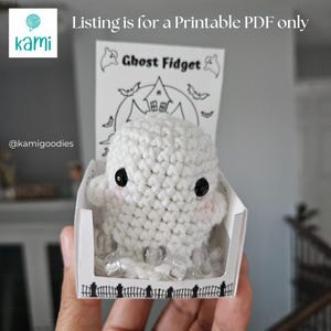 Könnte beinhalten: Ein weißes gehäkeltes Geister-Fidget-Spielzeug mit schwarzen Augen und rosa Wangen, präsentiert in einer dekorativen Box. Die Box hat ein Halloween-Design mit dem Text "Ghost Fidget". Das Angebot gilt für ein druckbares PDF.