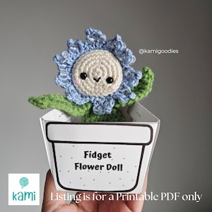 May include: A printable PDF pattern for a crochet Fidget Flower Doll. The doll is a blue flower with a white face and green leaves. The doll is sitting in a white pot with the text "Fidget Flower Doll" on it.