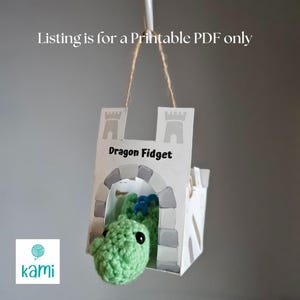 May include: A green crocheted dragon fidget toy peeking out of a white castle-shaped box. The box has gray accents and the words "Dragon Fidget" printed on it. The listing is for a printable PDF only.