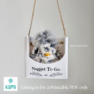 May include: A printable PDF pattern for a white and gray crocheted nugget toy with a black pom-pom. The toy is in a white box with a black handle and the text "Nugget-To-Go" printed on the front. The box is hanging from a string.
