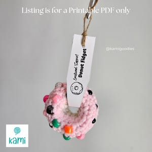 May include: A pink crocheted donut fidget toy with colorful beads and black buttons. The donut is hanging from a tag that says "Emotional Support Donut Fidget" and "@kamigoodies".