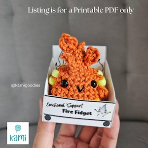 May include: An orange crochet fire-shaped fidget toy with black eyes and a smiling mouth, presented in a small box. The box reads "Emotional Support Fire Fidget" and "Listing is for a Printable PDF only."