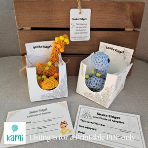 May include: Two crocheted snake fidget toys, one yellow and orange, the other blue, each in a decorative box. The boxes have the words "Snake Fidget" printed on them. Also included are adoption certificates with the text "Snake Fidget Certificate of Adoption".