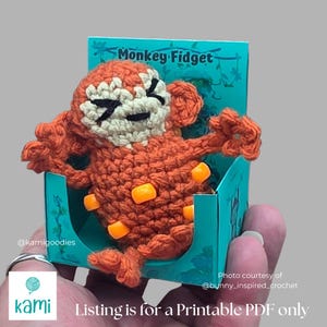 May include: A crocheted orange monkey fidget toy with a cream-colored face and black accents, presented in a teal box. The box reads "Monkey Fidget". The text "Listing is for a Printable PDF only" is also visible.