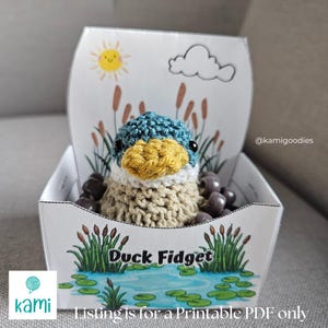 May include: A crocheted duck fidget toy with a blue and yellow head, nestled in a decorative box. The box features a pond scene with cattails, a smiling sun, and the text "Duck Fidget".