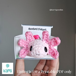 May include: A crocheted pink axolotl fidget toy with black eyes and a small pink heart on its head. The toy is displayed in a white and printed cardboard holder. The text "Axolotl Fidget" is printed on the holder.
