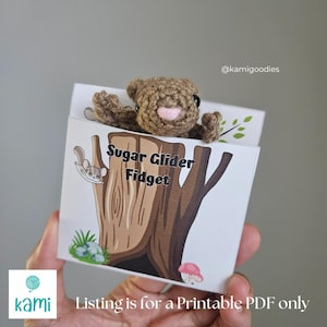 Może przedstawiać: Szydełkowa brązowa zabawka typu fidget z cukrowym szybowcem wystaje z białego pudełka. Pudełko przedstawia ilustrację pnia drzewa ze słowami "Sugar Glider Fidget". Mała ilustracja cukrowego szybowca znajduje się po lewej stronie.