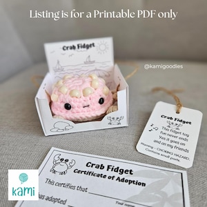 May include: A pink crocheted crab fidget toy with black eyes and cream-colored accents, presented in a decorative box. The box features a beach scene illustration and the text "Crab Fidget". A tag and adoption certificate are also included.