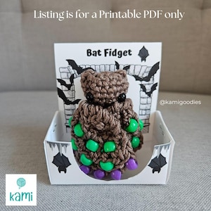 May include: A brown crocheted bat fidget toy with green and purple accents, displayed in a white box with a Halloween theme. The box features bat silhouettes and the text "Bat Fidget". The listing is for a printable PDF.