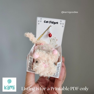 May include: A plush cat toy in a white box with the words "Cat Fidget". The toy is peach-colored with pink accents and a small red bell. The box has a cat illustration and the text "Listing is for a Printable PDF only".