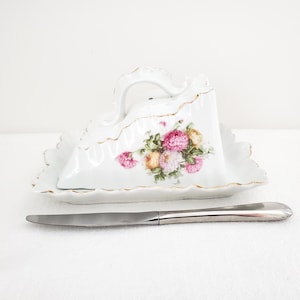 Peut inclure: Plat à fromage en porcelaine blanche avec un motif floral rose, jaune et pêche. Le plat a un bord festonné avec une bordure dorée et une poignée assortie. Un couteau en acier inoxydable repose devant.