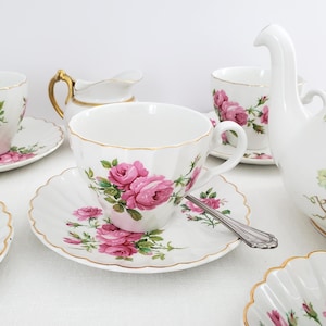 Może przedstawiać: Biały porcelanowy zestaw filiżanki i spodka z różowym wzorem róż i złotymi obwódkami. Srebrna łyżeczka spoczywa na spodku. Inne elementy to śmietanka i czajniczek, wszystkie z pasującymi wzorami róż.